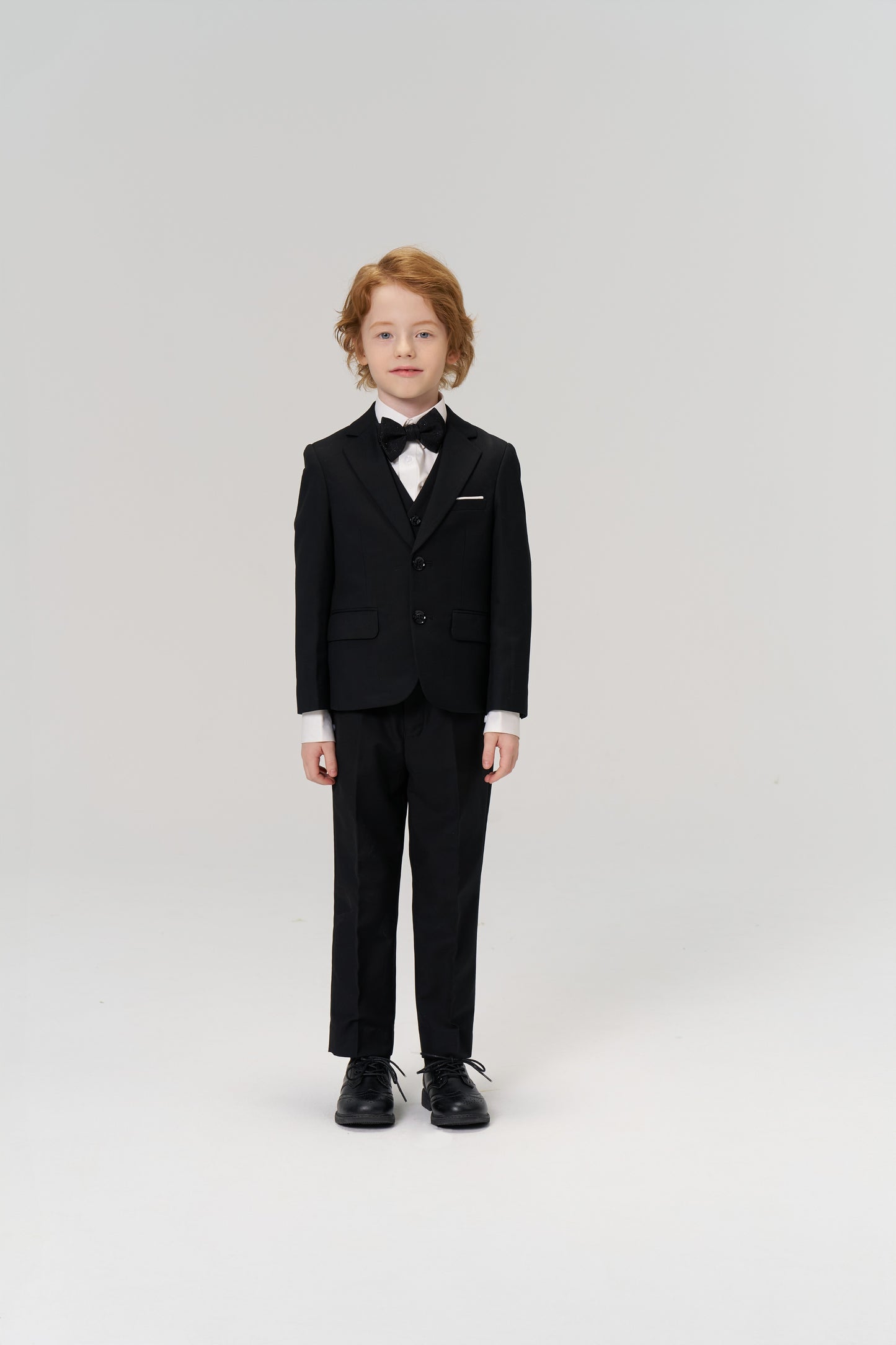 A young boy with light hair faces forward in KIKI and JOJO's Noir Bloom Suit Pants, styled with a black suit jacket, bow tie, and dress shoes—an ideal choice for formal occasions—set against a plain white background.