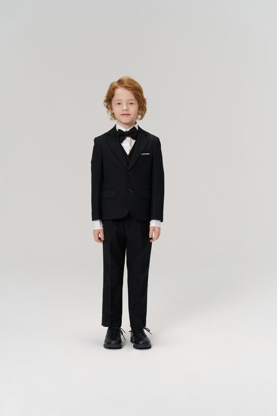 A young boy with light hair faces forward in KIKI and JOJO's Noir Bloom Suit Pants, styled with a black suit jacket, bow tie, and dress shoes—an ideal choice for formal occasions—set against a plain white background.