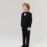 A young boy with light brown hair wears KIKI and JOJO's Noir Bloom Suit Pants, styled with a black suit jacket, white shirt, and bow tie against a plain light background—ideal for formal occasions.