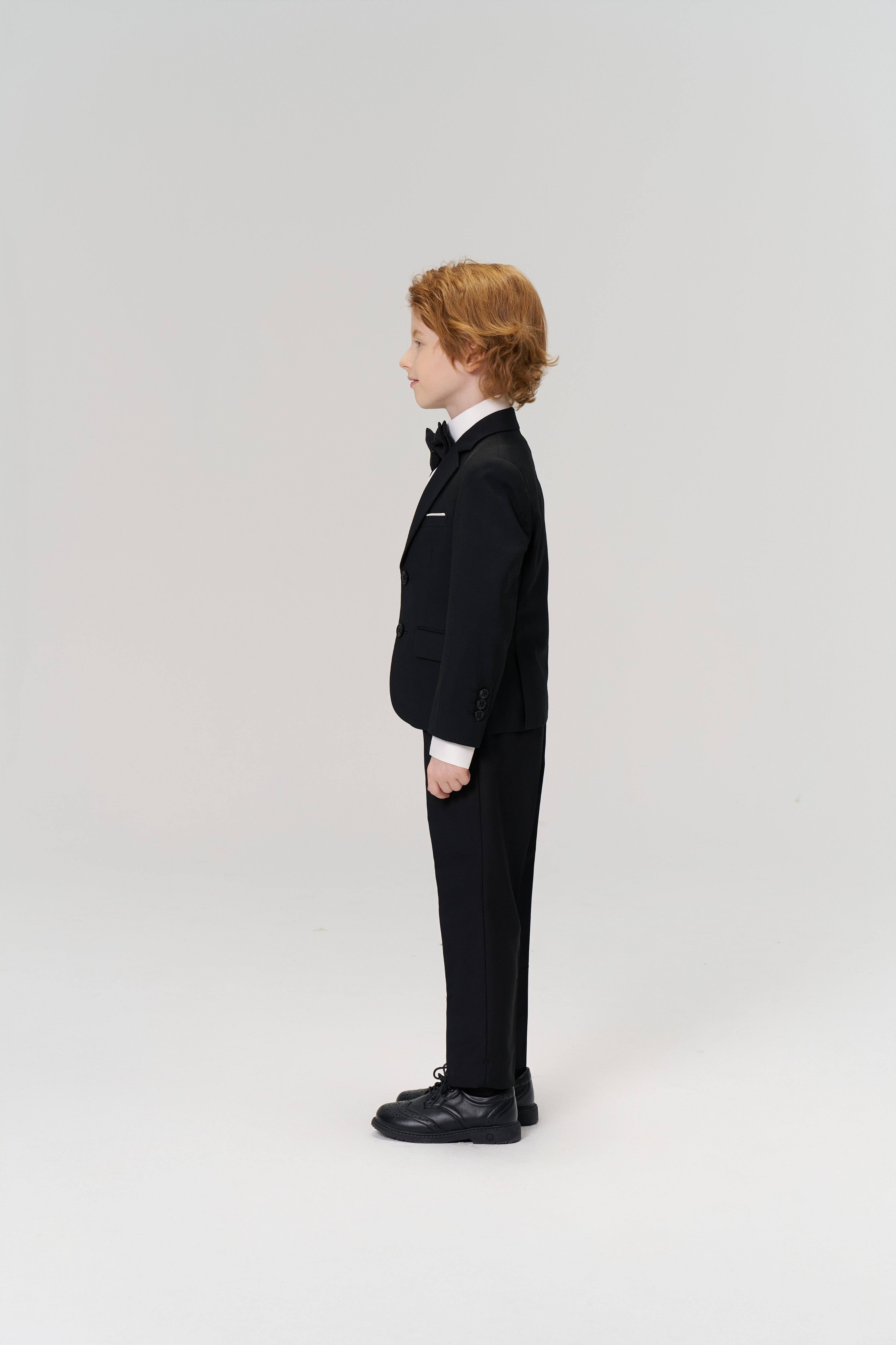 A young boy with light brown hair stands in profile, wearing KIKI and JOJO Noir Bloom Suit Pants—sleek black straight-leg trousers—paired with a tuxedo and bow tie, perfect for formal occasions, against a plain white background.