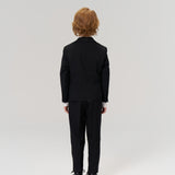 A young child with light brown hair, dressed in KIKI and JOJO's Noir Bloom Suit Pants, stands facing away from the camera against a plain light background—classic attire for formal occasions.