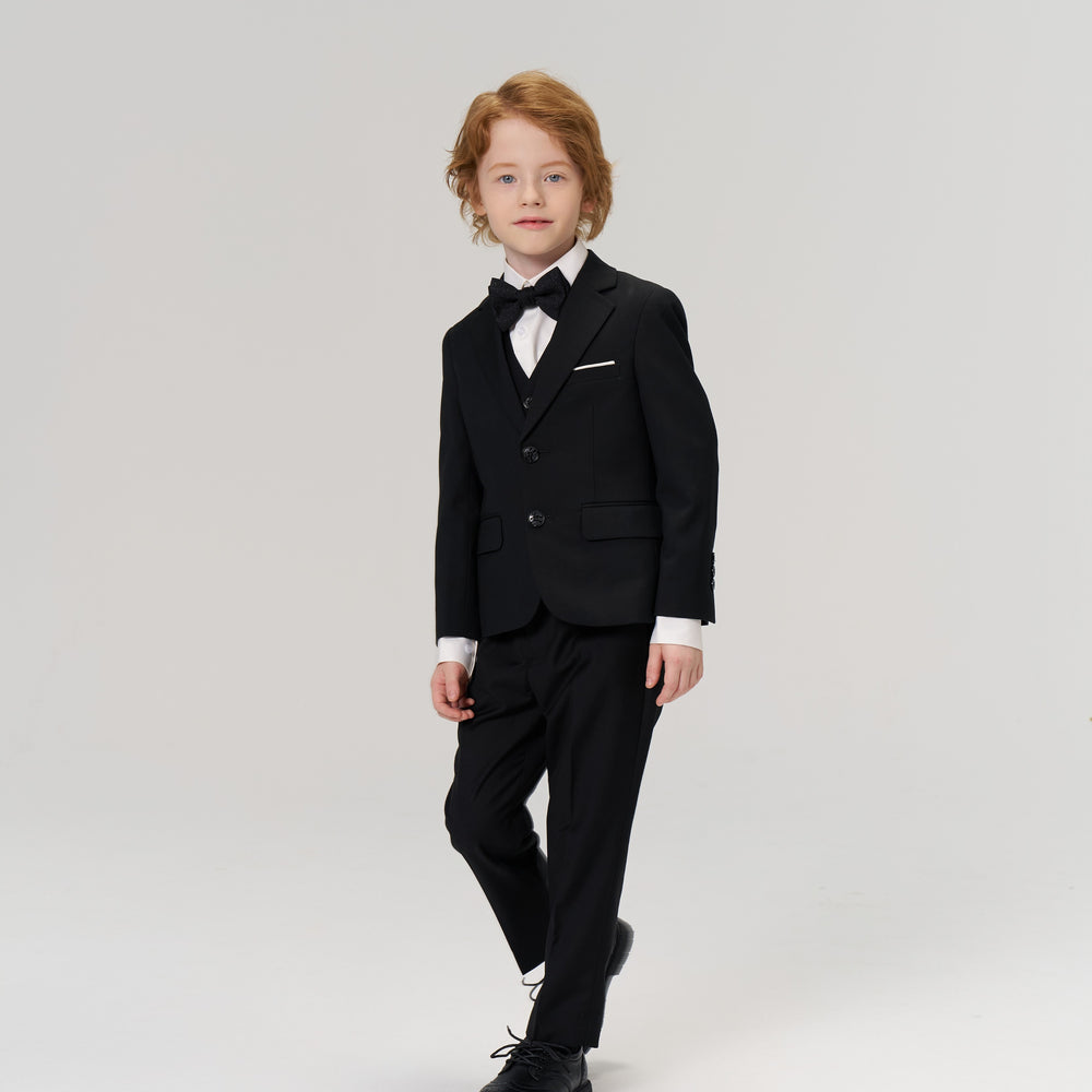 A young boy with light brown hair is dressed in a black suit and bow tie, standing against a plain, light background.