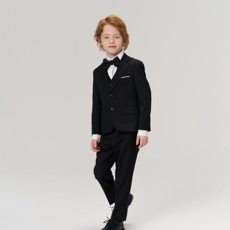 A young boy with light brown hair is dressed in a black suit and bow tie, standing against a plain, light background.