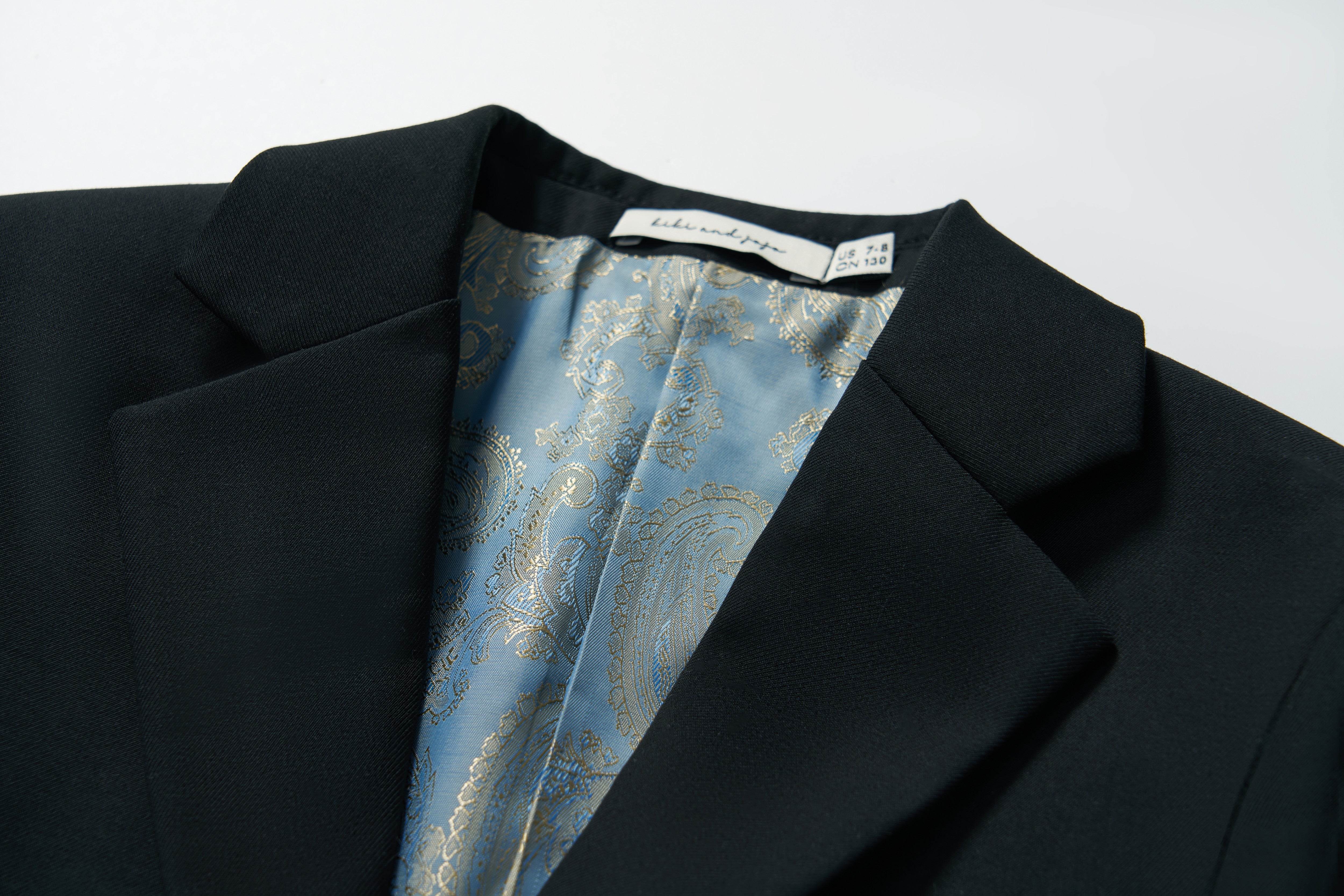A close-up of the Noir Bloom Suit Jacket by KIKI and JOJO, a boys' black jacket with a classic notched lapel, blue paisley-patterned lining, and a visible brand label sewn into the collar area.