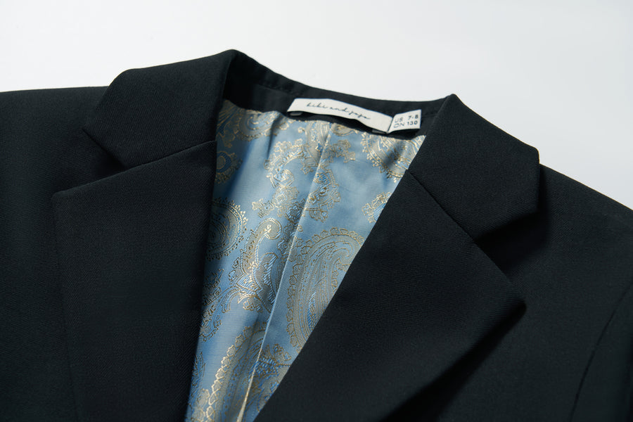 A close-up of the Noir Bloom Suit Jacket by KIKI and JOJO, a boys' black jacket with a classic notched lapel, blue paisley-patterned lining, and a visible brand label sewn into the collar area.