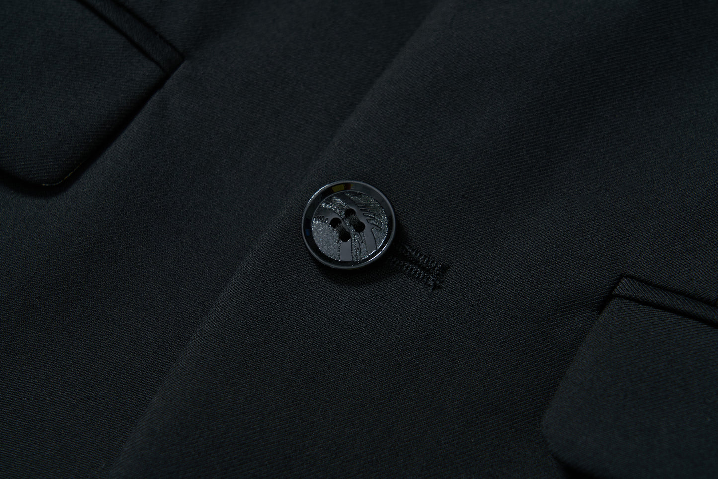 Close-up of the KIKI and JOJO Noir Bloom Suit Jacket shows a fastened button on black fabric, classic notched lapel, fine stitching, and two pocket flaps on each side.