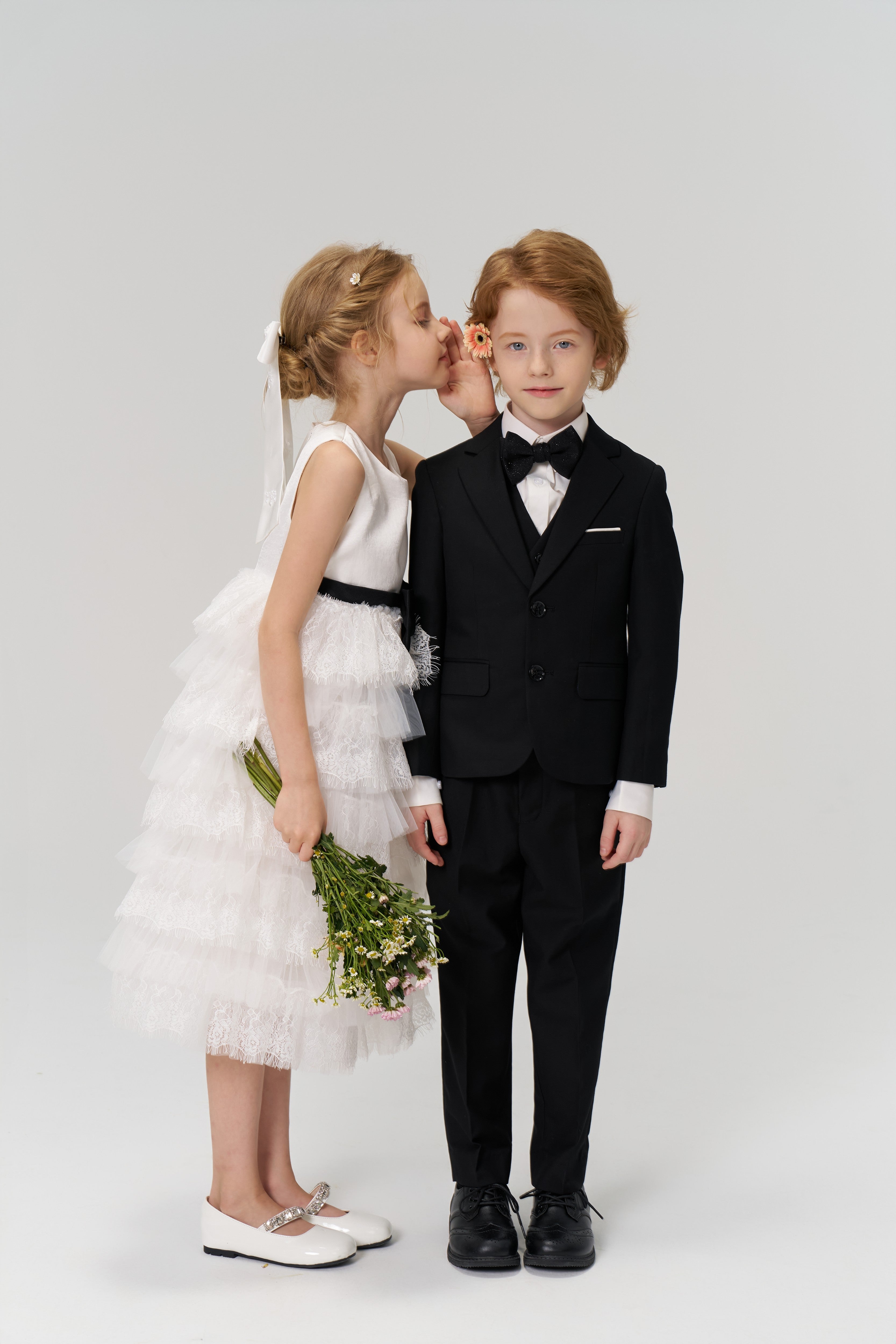 A girl whispering to a boy in a tuxedo.