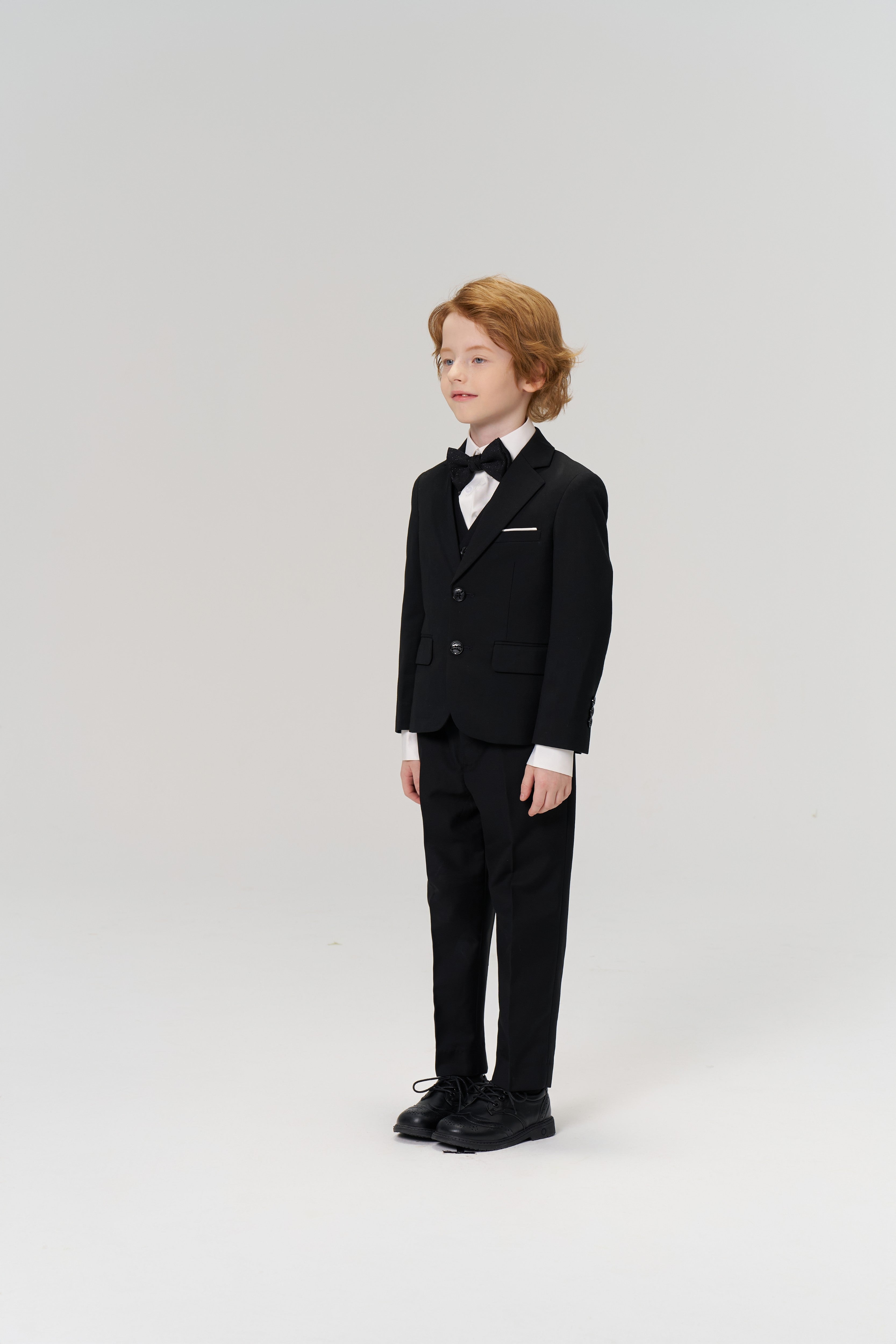 A young boy with light brown hair stands against a plain light background wearing the KIKI and JOJO Noir Bloom Suit Jacket, featuring a classic notched lapel, paired with a white shirt and black bow tie.