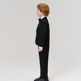 A young boy in a KIKI and JOJO Noir Bloom Suit Jacket with classic notched lapels and a bow tie stands in profile against a plain light background.