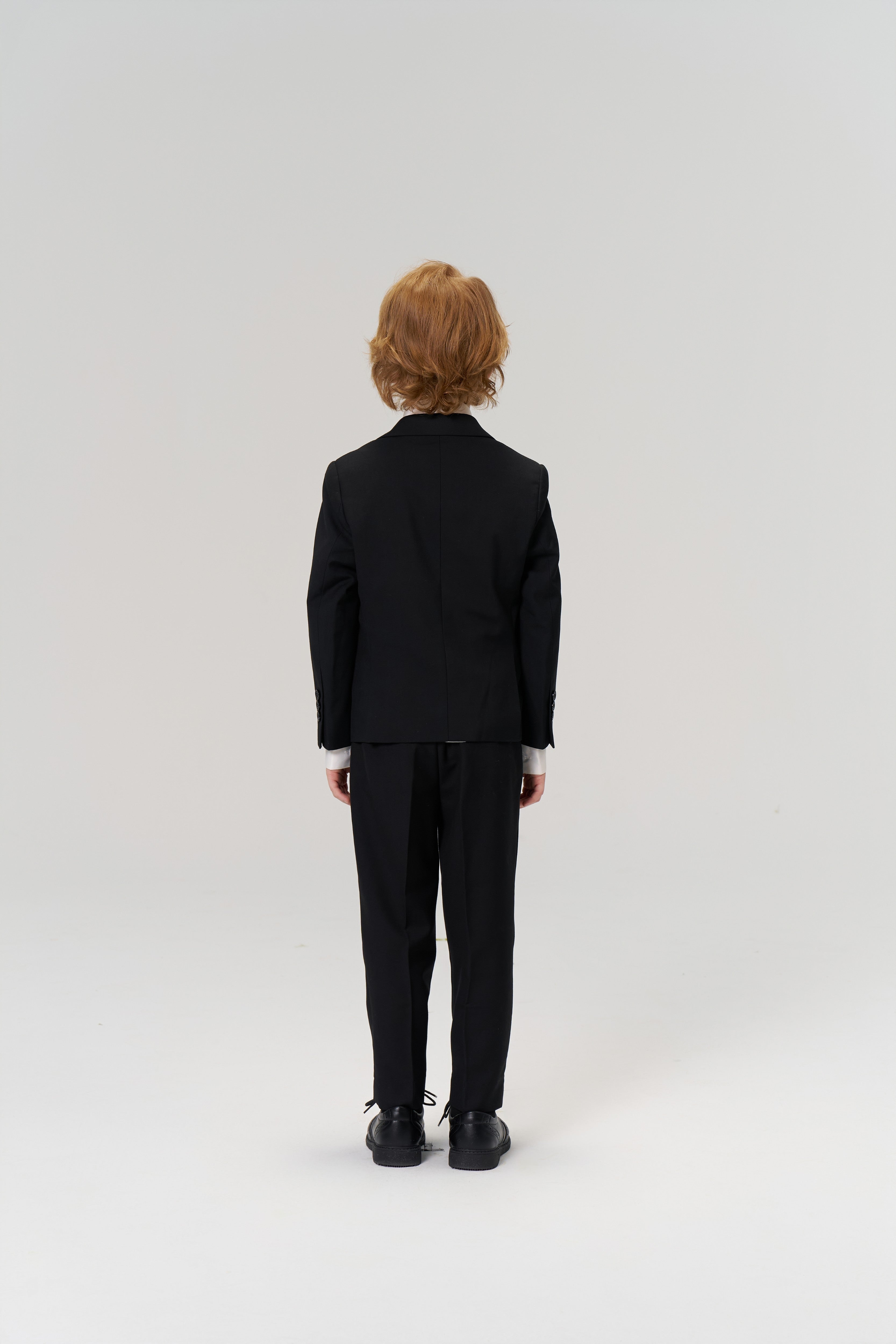 A young child with light brown hair, wearing the KIKI and JOJO Noir Bloom Suit Jacket and black shoes, stands facing away from the camera against a plain, light background.
