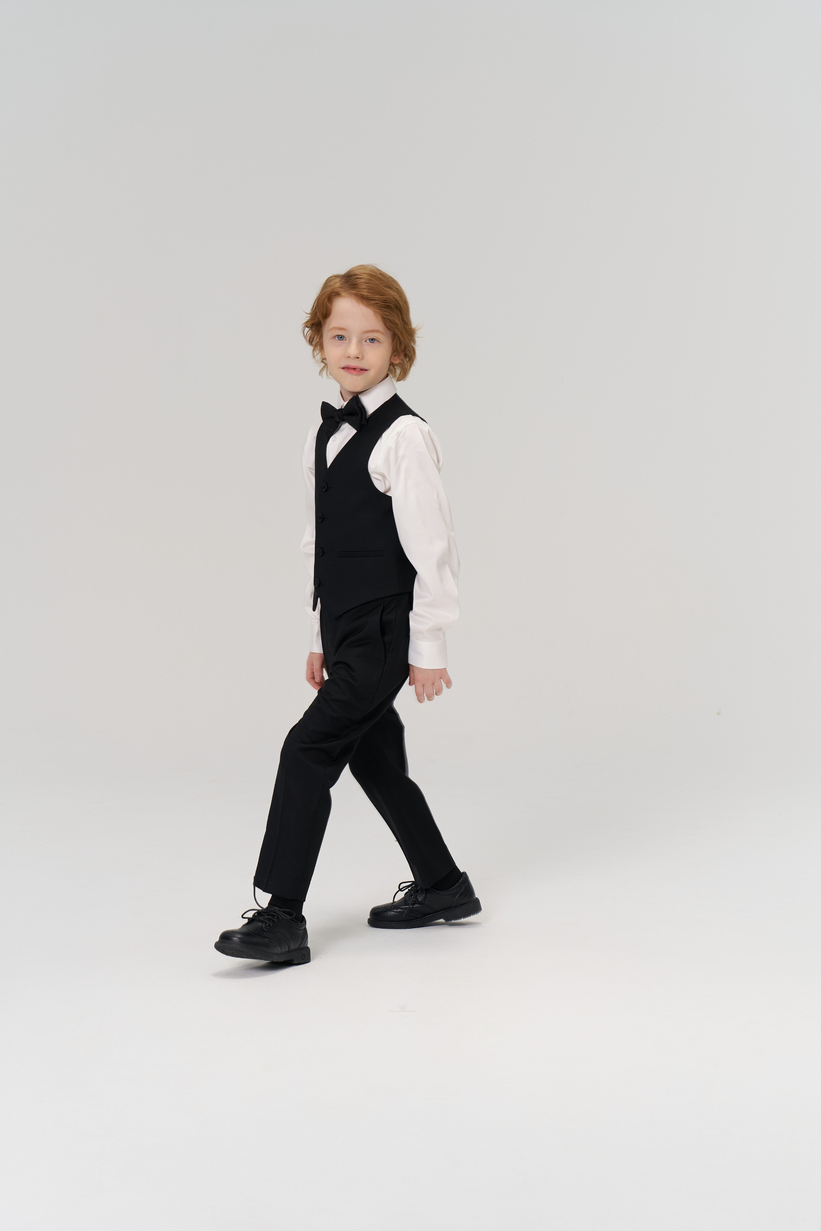 A young boy with shoulder-length hair wears the KIKI and JOJO Noir Bloom Suit Vest, styled with a bow tie and dress shoes, as he walks against a plain white background.