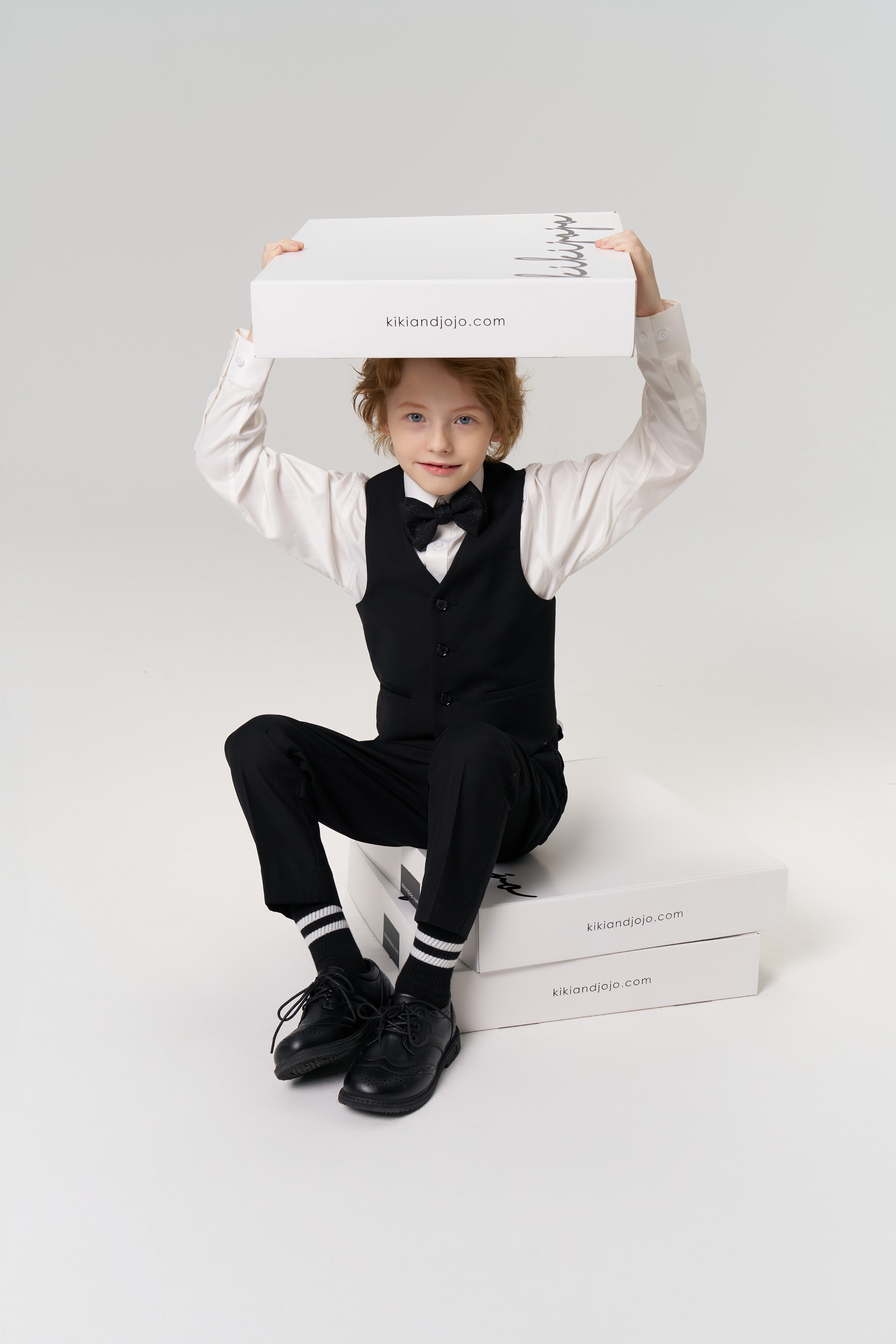 A boy in a tailored Noir Bloom Suit Vest by KIKI and JOJO, paired with a bow tie, sits on white boxes labeled "kikilandjojo.com," holding another box overhead—a classic touch for any formal wardrobe.