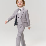 A young boy with light brown hair wears a light gray suit, bow tie, and black shoes, walking confidently against a plain light background.
