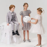 Three children dressed in formal clothes interact; two girls in white and gray dresses, and a boy in a gray suit sit and stand against a plain background, one girl holding a white balloon.