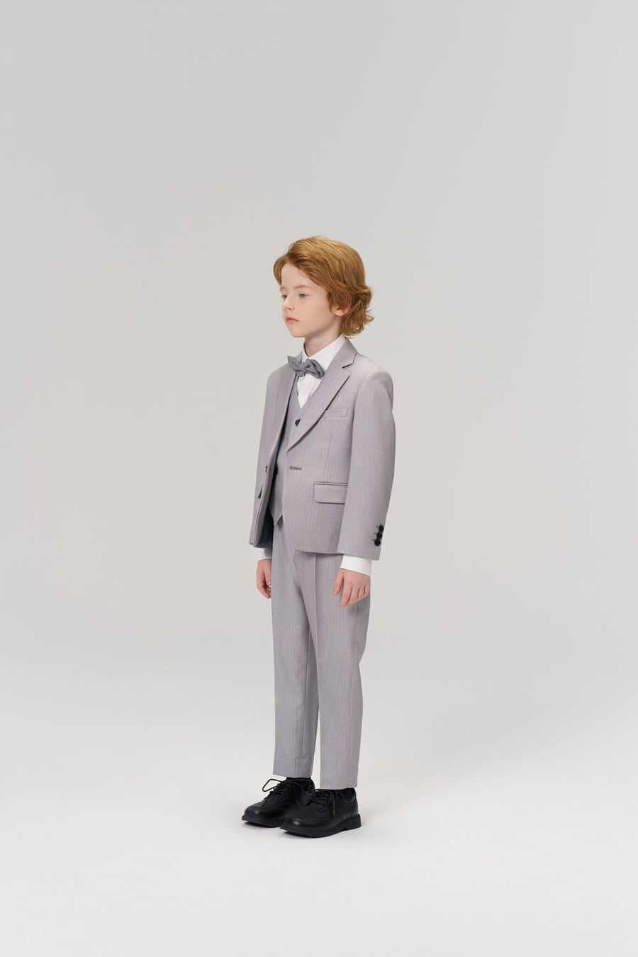 A young boy wears KIKI and JOJO’s Silver Bloom Suit Pants with an adjustable waistband for comfort, paired with a light gray pinstripe suit, bow tie, and black shoes, as he stands against a plain light background facing slightly left.