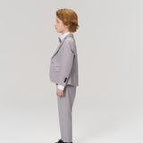 A young boy with light brown hair stands in profile, wearing KIKI and JOJO Silver Bloom Suit Pants with an adjustable waistband, a light gray suit, white shirt, bow tie, and black shoes against a plain light background.