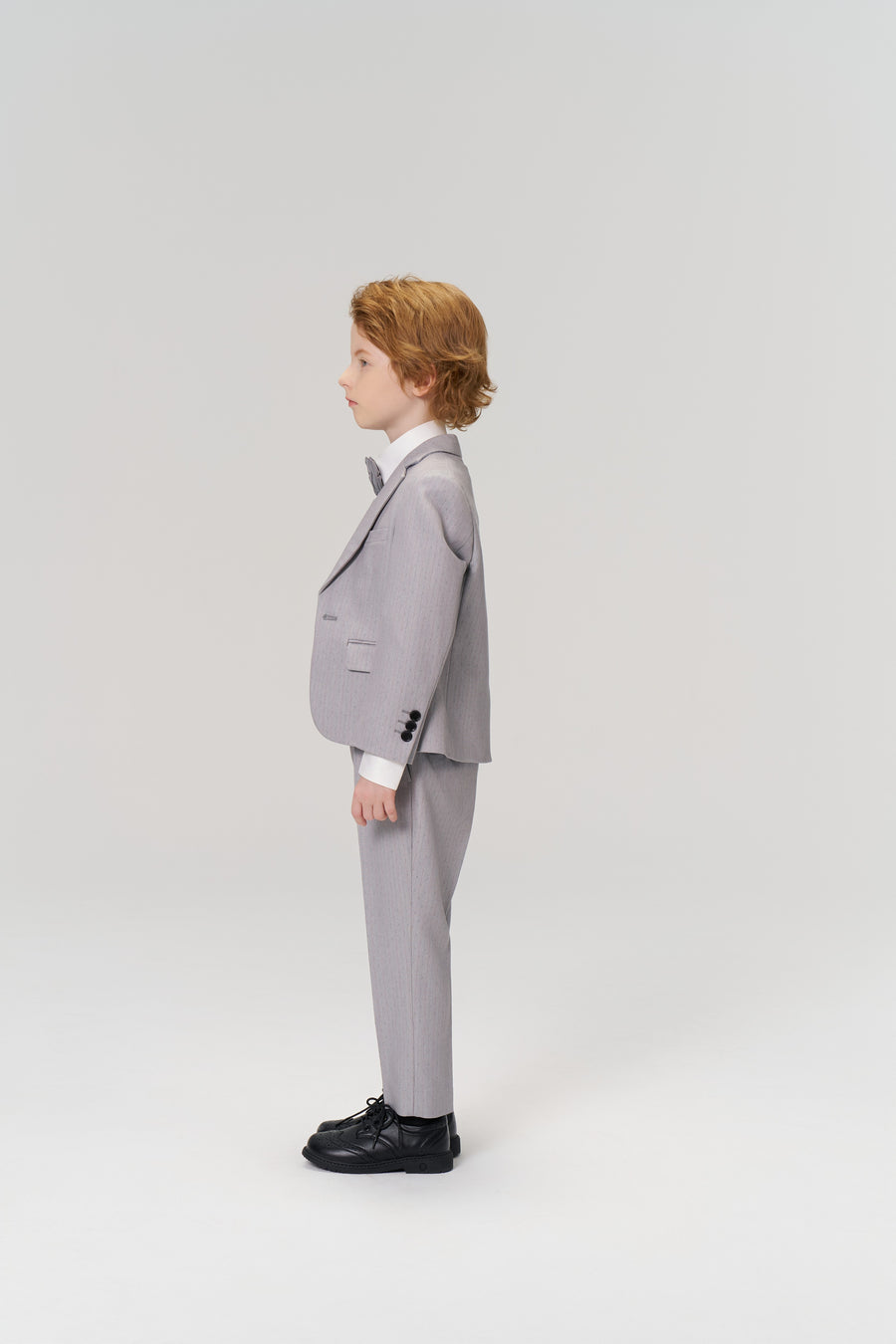 A young boy with light brown hair stands in profile, wearing KIKI and JOJO Silver Bloom Suit Pants with an adjustable waistband, a light gray suit, white shirt, bow tie, and black shoes against a plain light background.