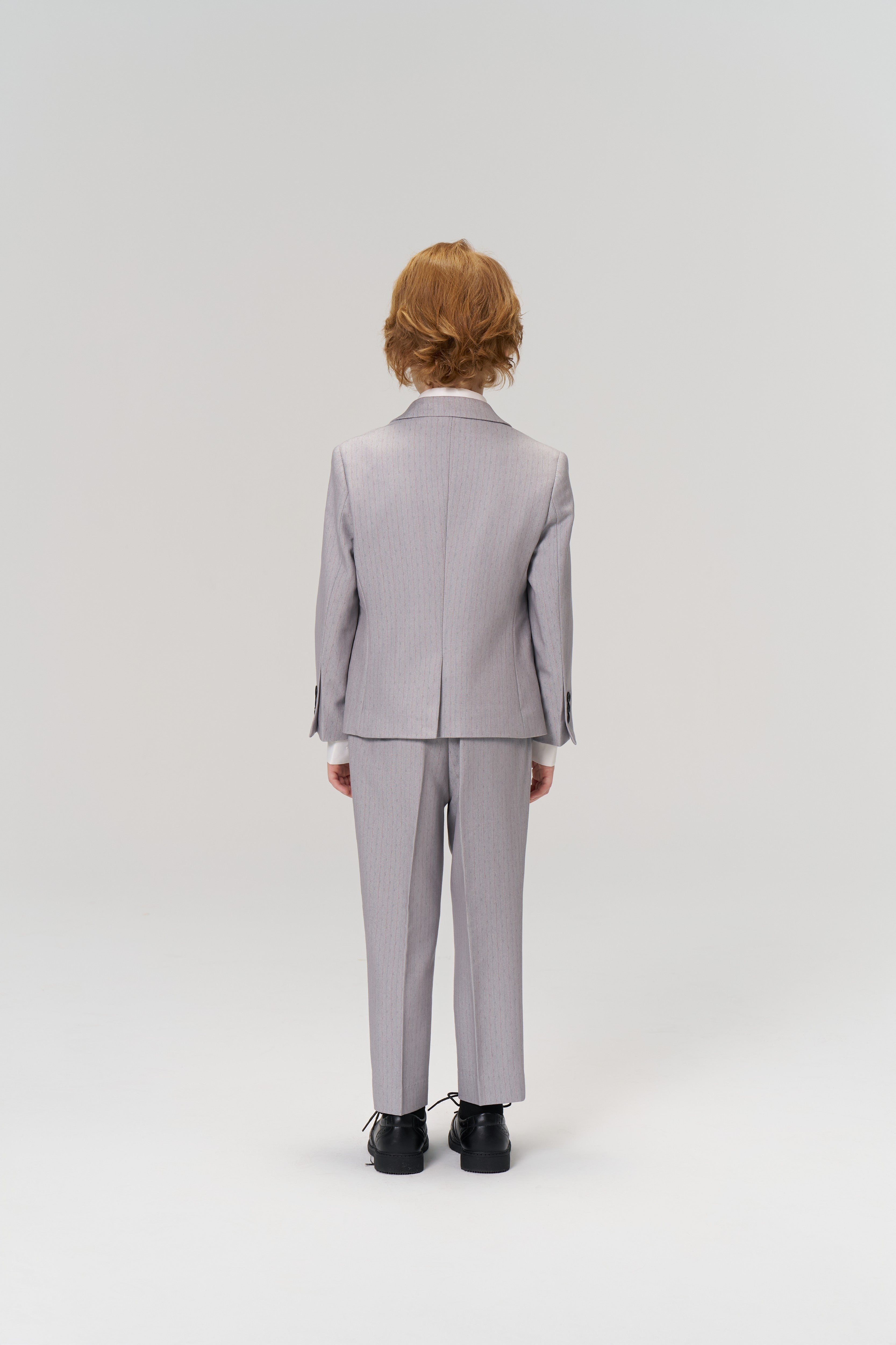 A young person with light brown hair stands facing away in KIKI and JOJO’s Silver Bloom Suit Pants, featuring an adjustable waistband, paired with black shoes against a plain white background.