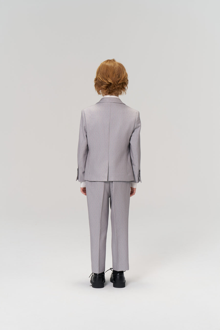 A young person with light brown hair stands facing away in KIKI and JOJO’s Silver Bloom Suit Pants, featuring an adjustable waistband, paired with black shoes against a plain white background.