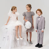 Three children in formal attire pose against a plain background; two girls wear dresses, and one boy wears a light gray suit with a bow tie.