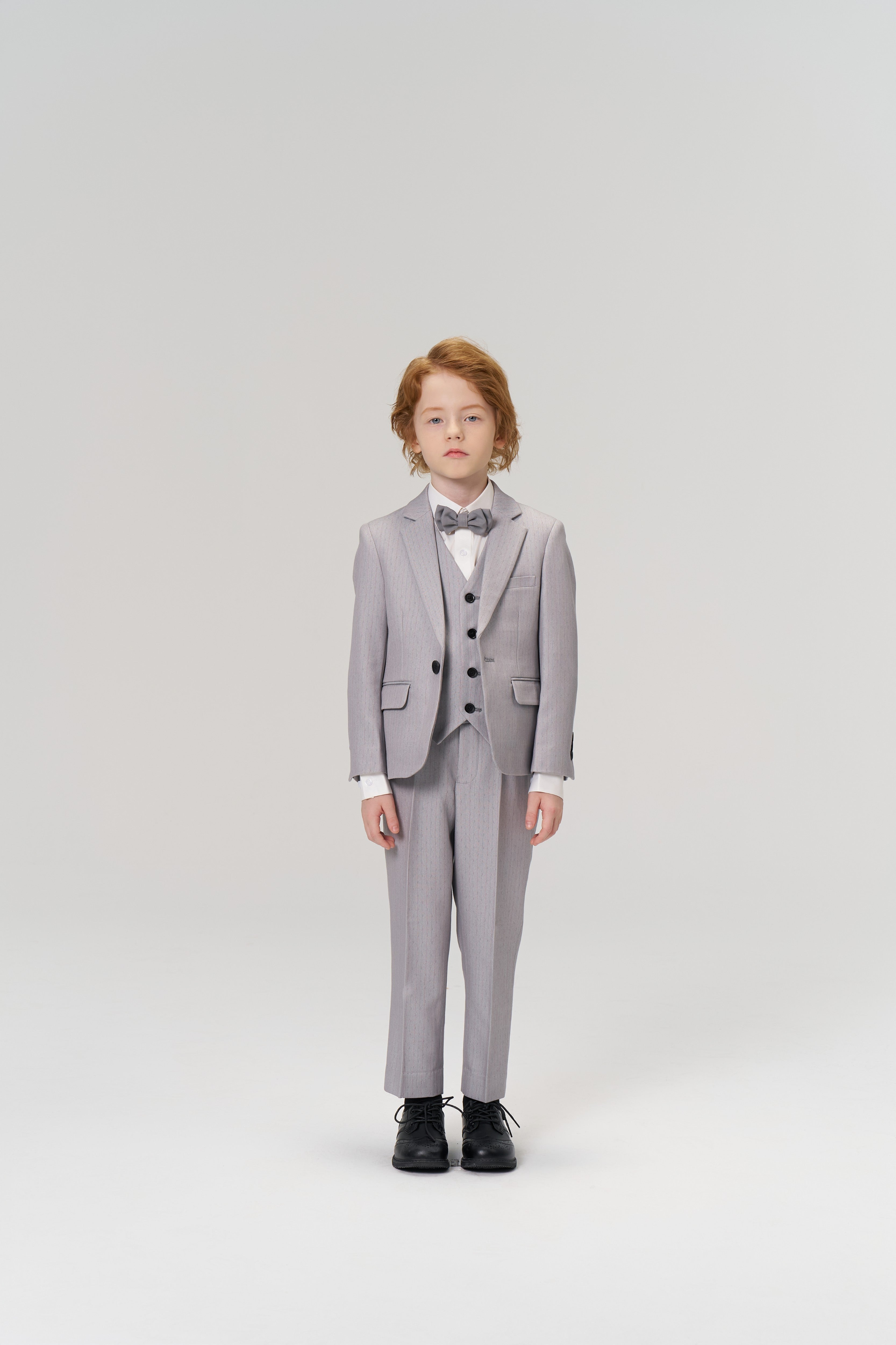 A young child with light brown hair wears the KIKI and JOJO Silver Bloom Suit Jacket, featuring light gray stripes, a bow tie, and black shoes against a plain studio background—ideal for formal boys’ occasions.