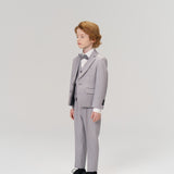 A young boy with light brown hair stands in the KIKI and JOJO Silver Bloom Suit Jacket over a white shirt and bow tie, set against a plain, light background.