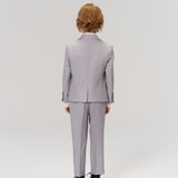 A child with light brown hair, wearing the KIKI and JOJO Silver Bloom Suit Jacket and black shoes, faces away from the camera against a plain, light background.