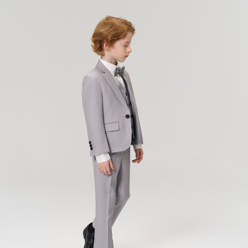 A young boy with light brown hair, dressed in a light gray suit with a bow tie and black shoes, stands against a plain, light background.