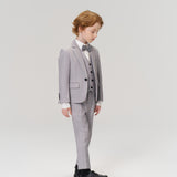 A young boy with light brown hair looks down while wearing the KIKI and JOJO Silver Bloom Suit Vest, a tailored light grey suit, bow tie, and black shoes against a plain light background.