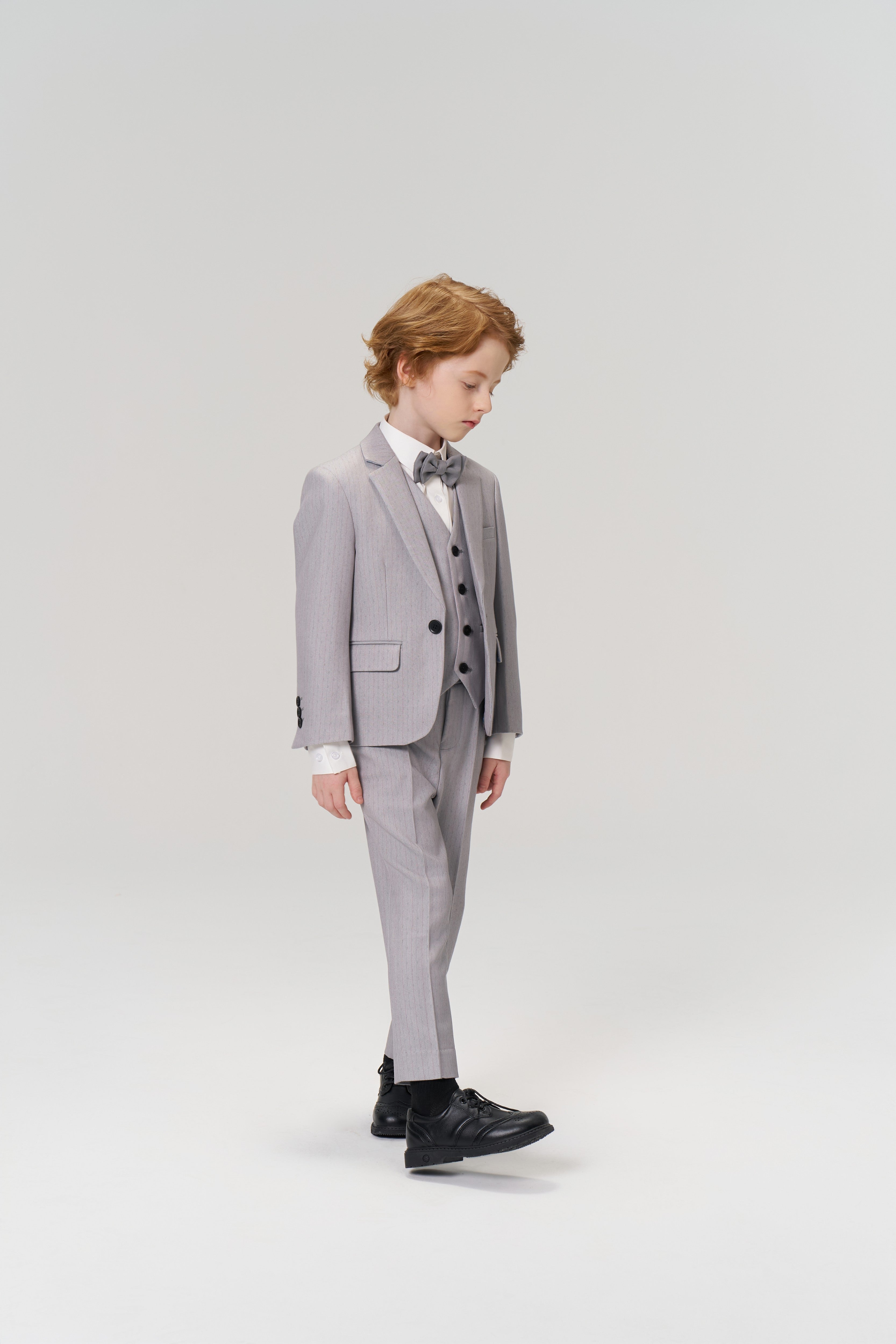 A young boy with light brown hair looks down while wearing the KIKI and JOJO Silver Bloom Suit Vest, a tailored light grey suit, bow tie, and black shoes against a plain light background.
