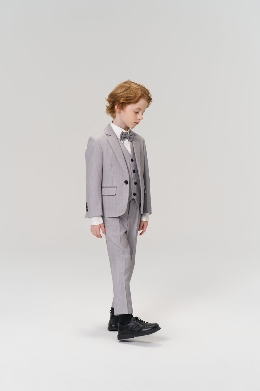 A young boy with light brown hair looks down while wearing the KIKI and JOJO Silver Bloom Suit Vest, a tailored light grey suit, bow tie, and black shoes against a plain light background.
