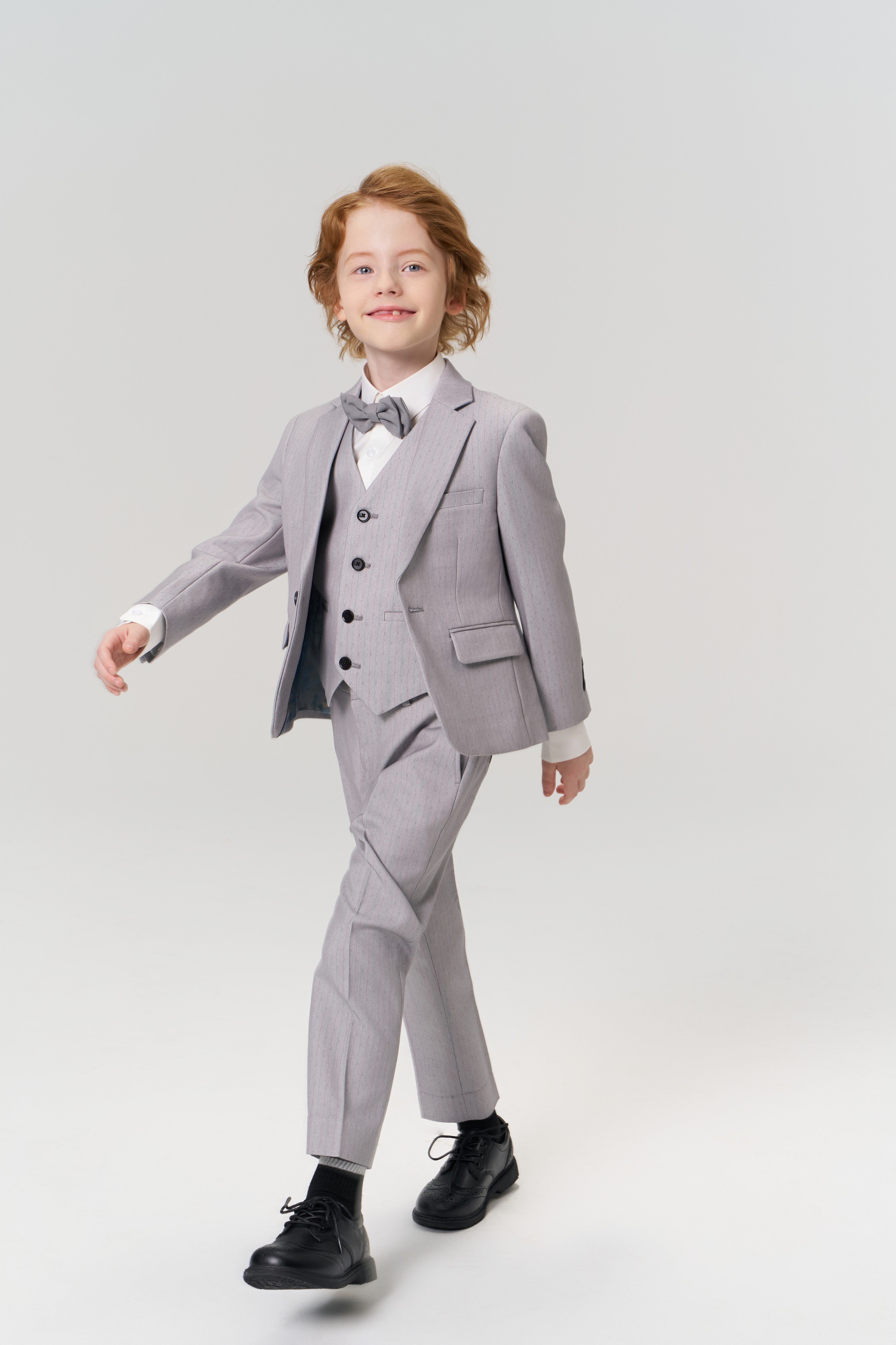 A young boy with light brown hair models the KIKI and JOJO Silver Bloom Suit Vest, styled with a light gray suit, white shirt, and bow tie, as he walks forward against a plain light background.
