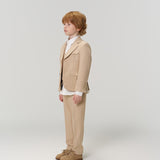 A young boy with light brown hair stands in profile wearing KIKI and JOJO's Savoy Dune Suit Pants with an adjustable waistband, a white shirt, and beige shoes against a plain light background.