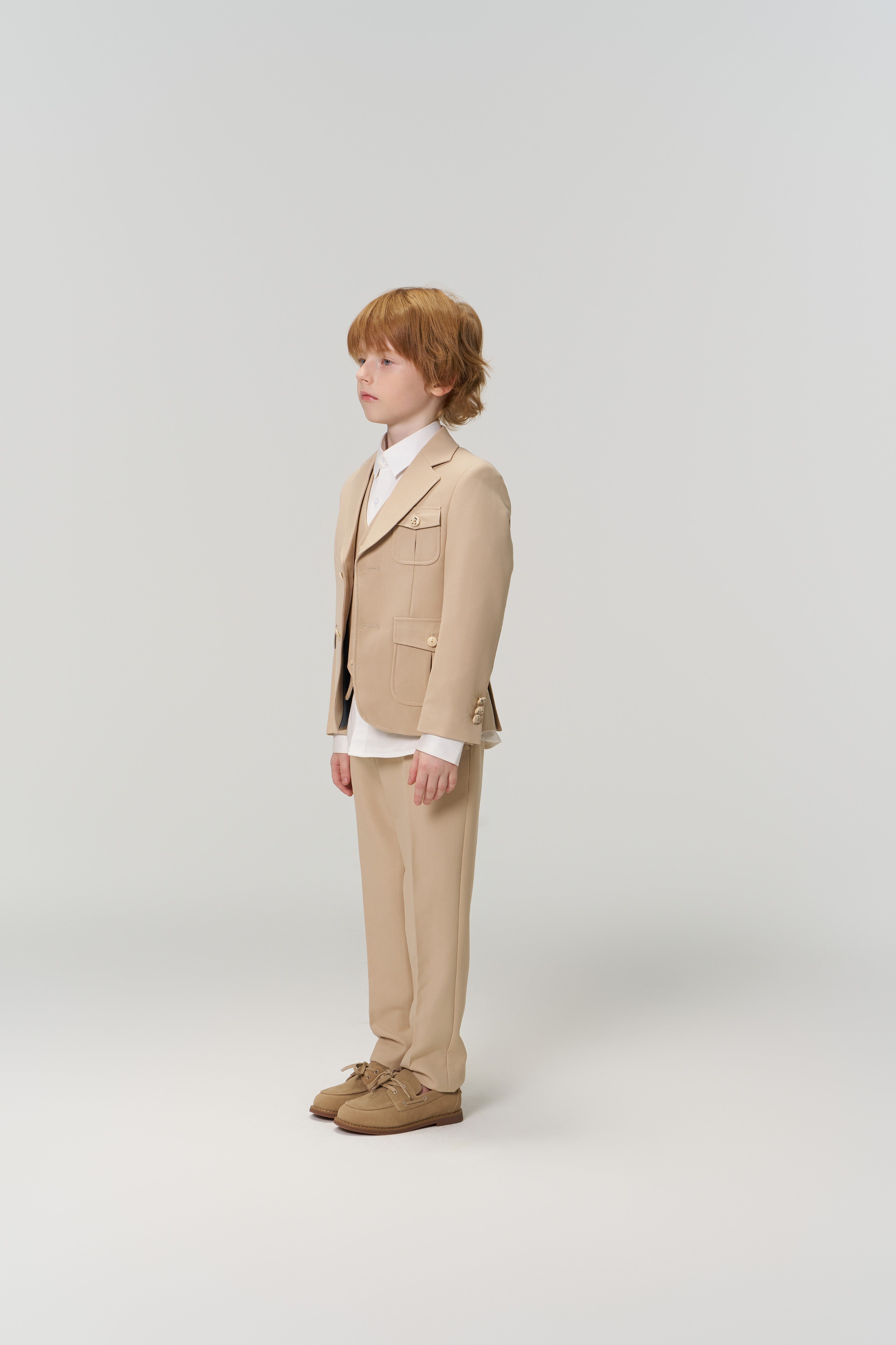A young boy with light brown hair stands in profile wearing KIKI and JOJO's Savoy Dune Suit Pants with an adjustable waistband, a white shirt, and beige shoes against a plain light background.