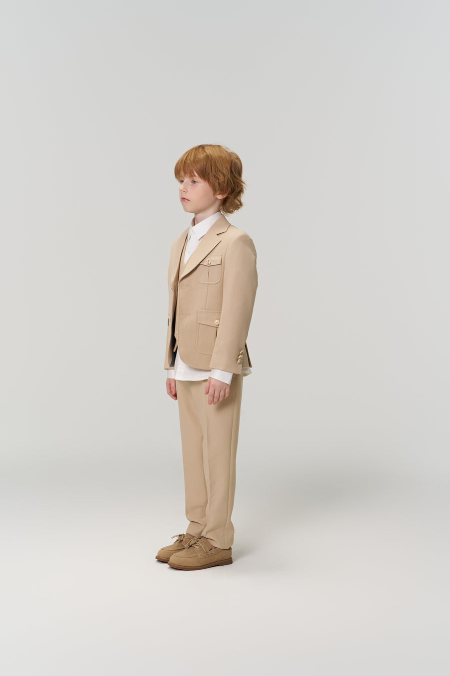 A young boy with light brown hair stands in profile wearing KIKI and JOJO's Savoy Dune Suit Pants with an adjustable waistband, a white shirt, and beige shoes against a plain light background.