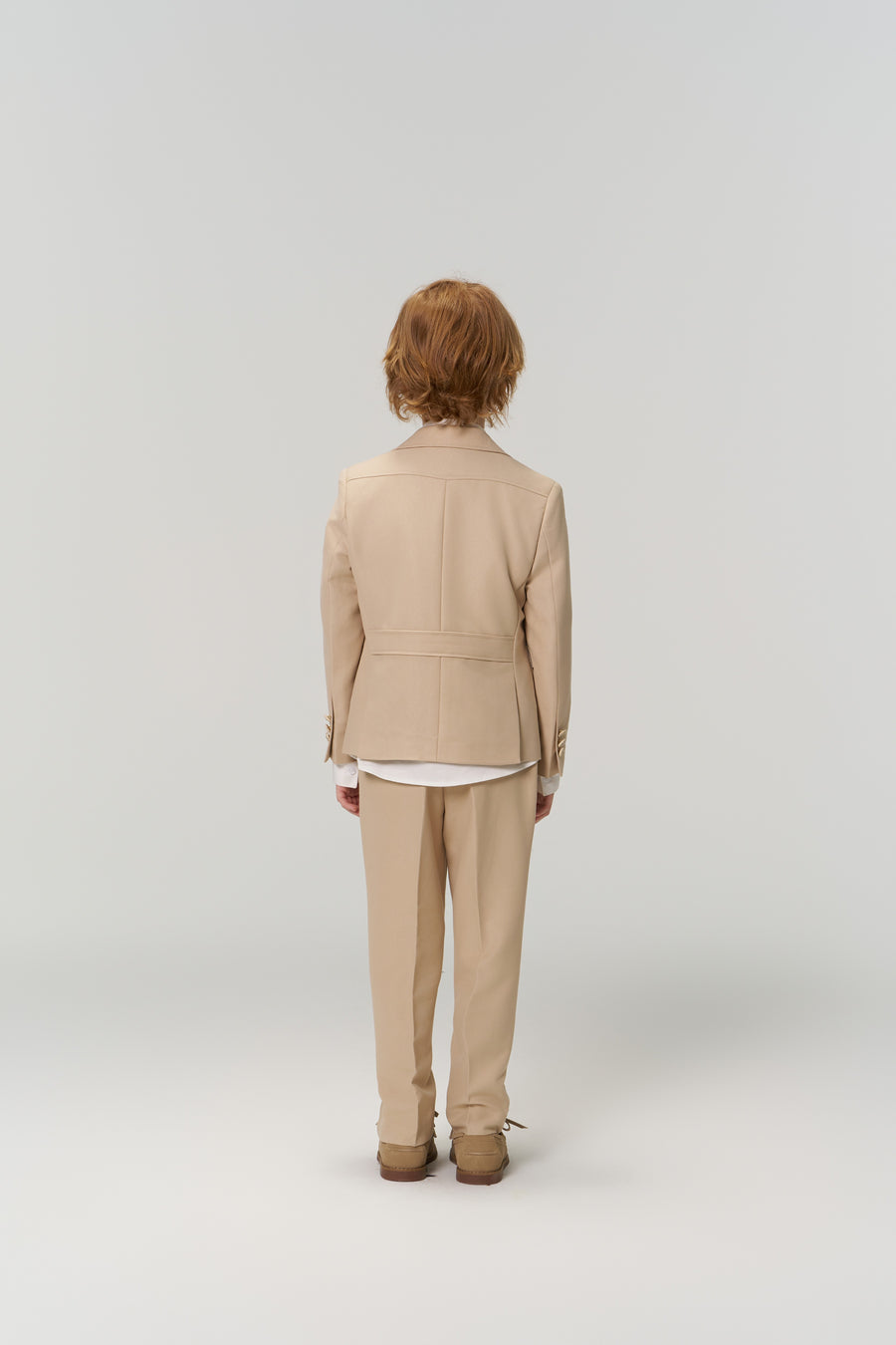 A person with short, light brown hair in a beige suit faces away from the camera against a plain background, showcasing KIKI and JOJO’s Savoy Dune Suit Pants for boys, featuring an adjustable waistband for comfort.