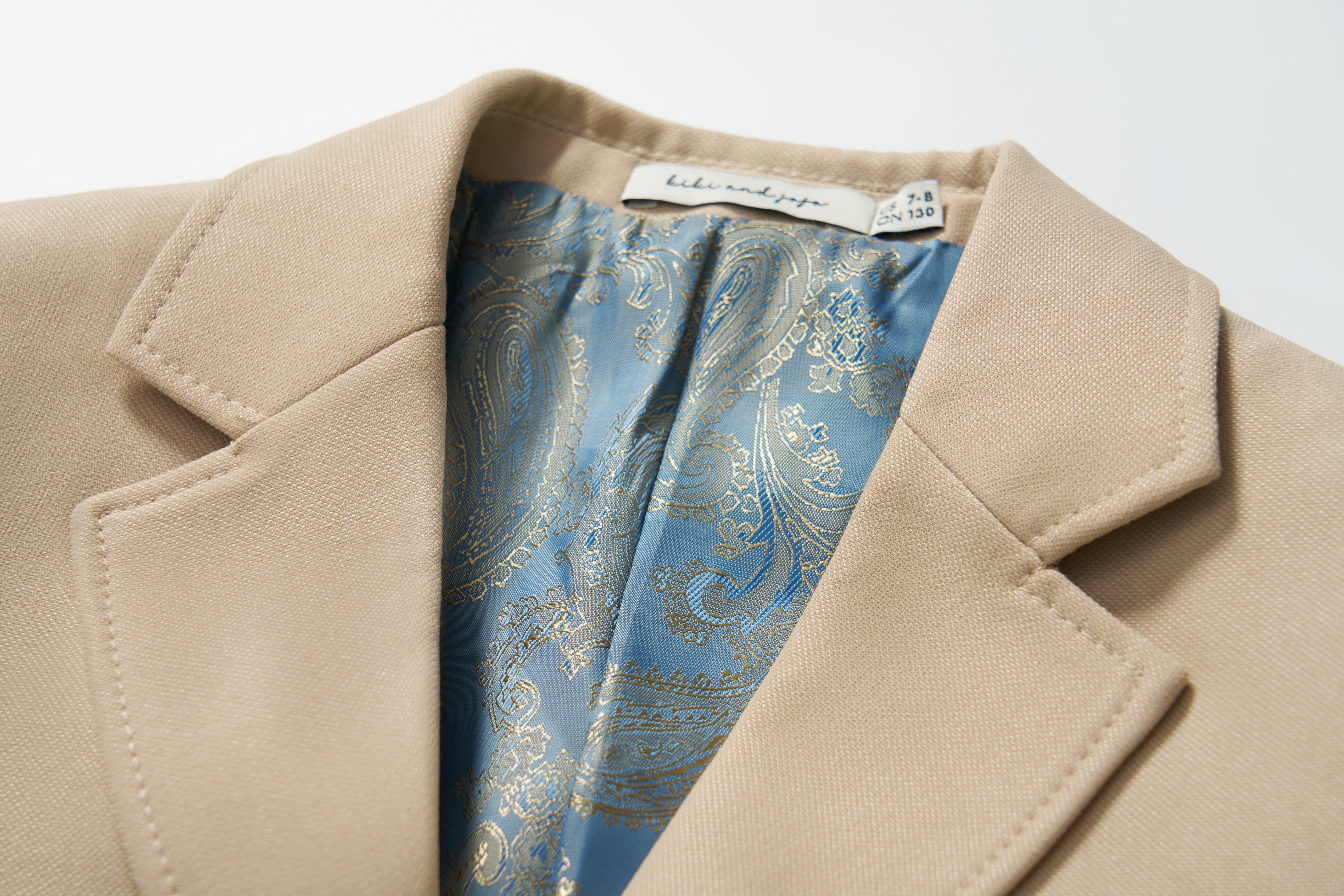 Close-up of the KIKI and JOJO Savoy Dune Suit Jacket in beige, featuring classic tailoring, a blue and gold paisley patterned lining, and a visible neckline label.