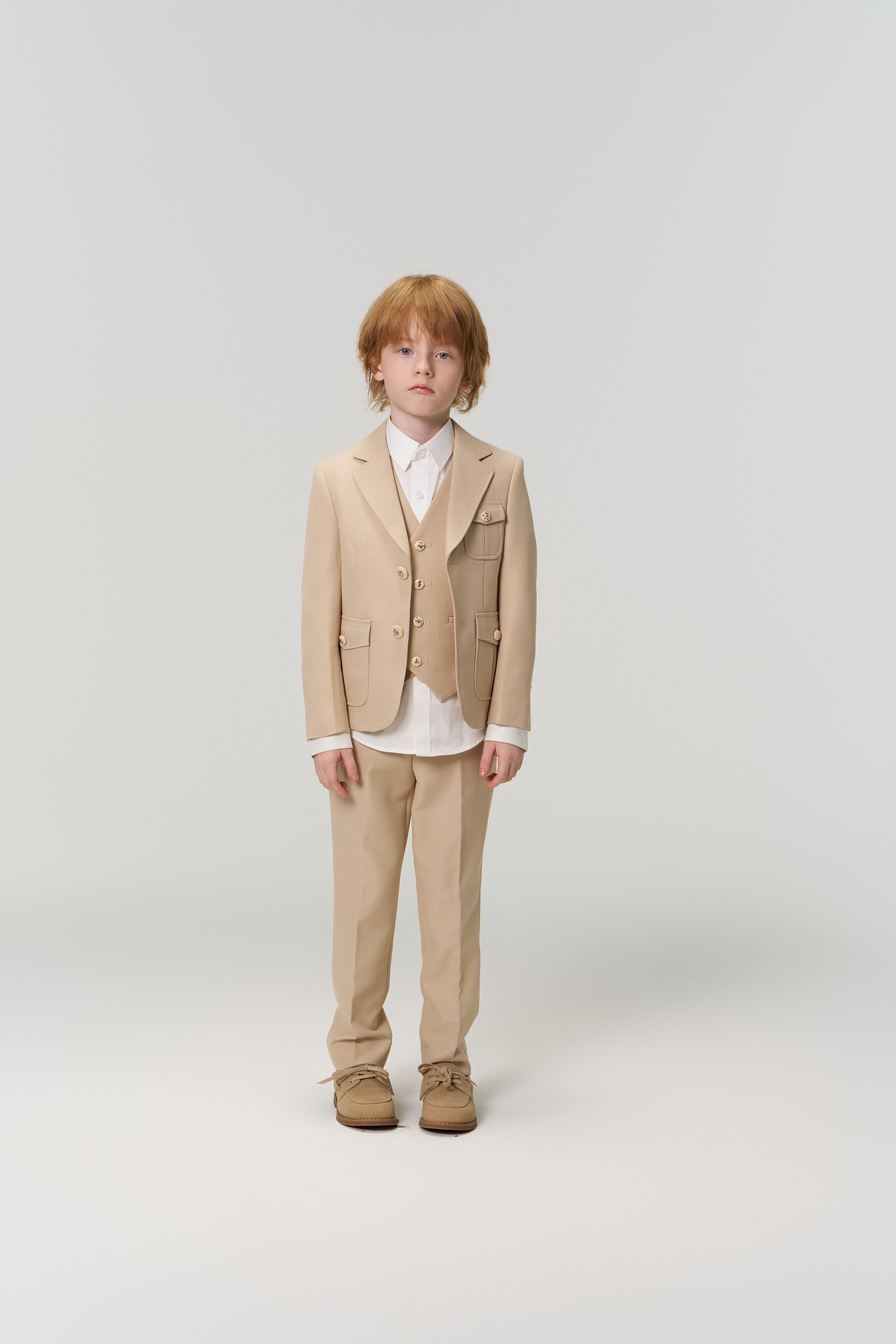 A young boy with light brown hair faces forward, wearing the KIKI and JOJO Savoy Dune Suit Jacket over a white shirt and brown shoes, exemplifying classic boys’ formalwear against a plain light background.