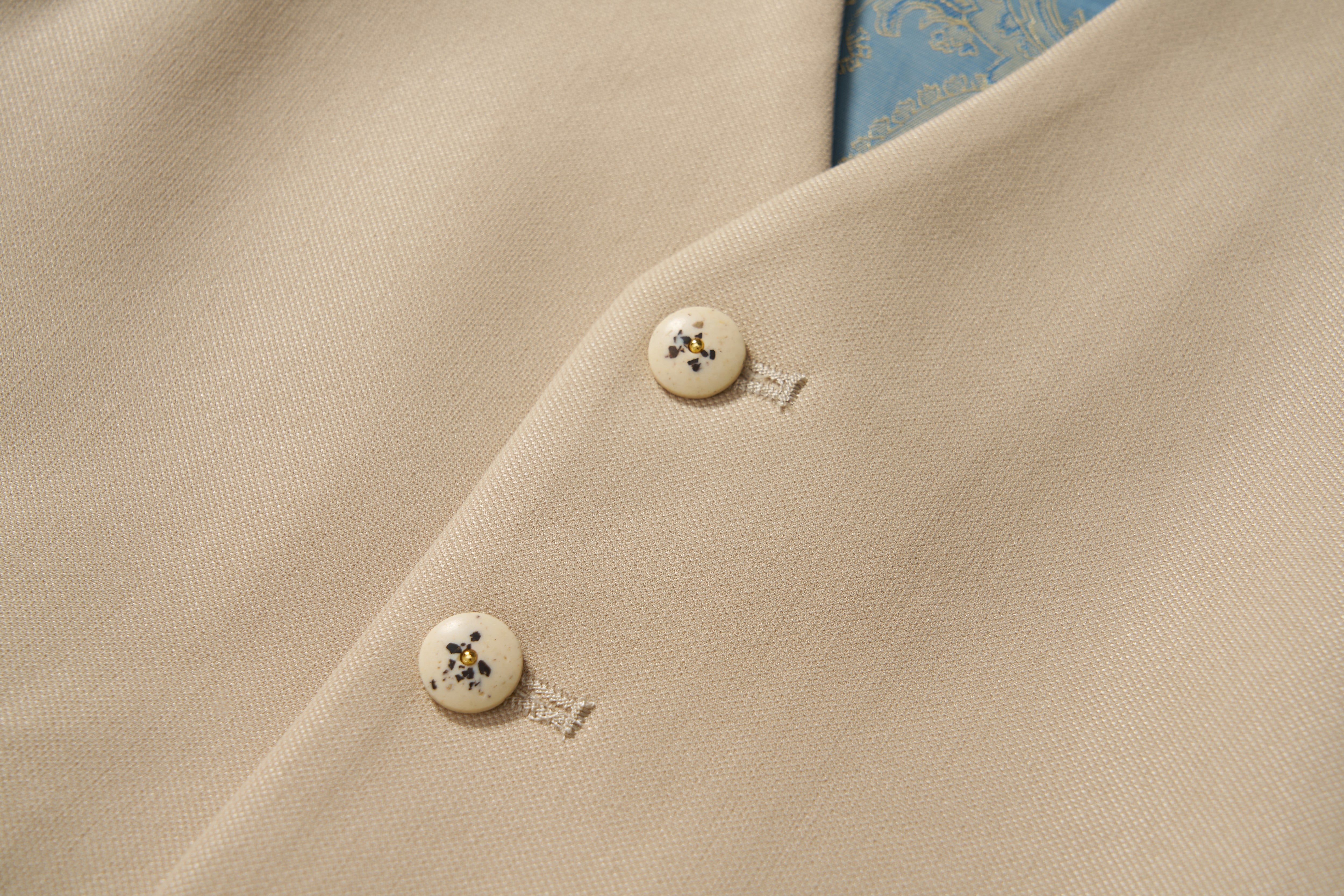 Close-up of a khaki beige blazer with cream decorative buttons featuring a black floral pattern and blue paisley lining—an ideal match for the KIKI and JOJO Savoy Dune Suit Vest on any special occasion.