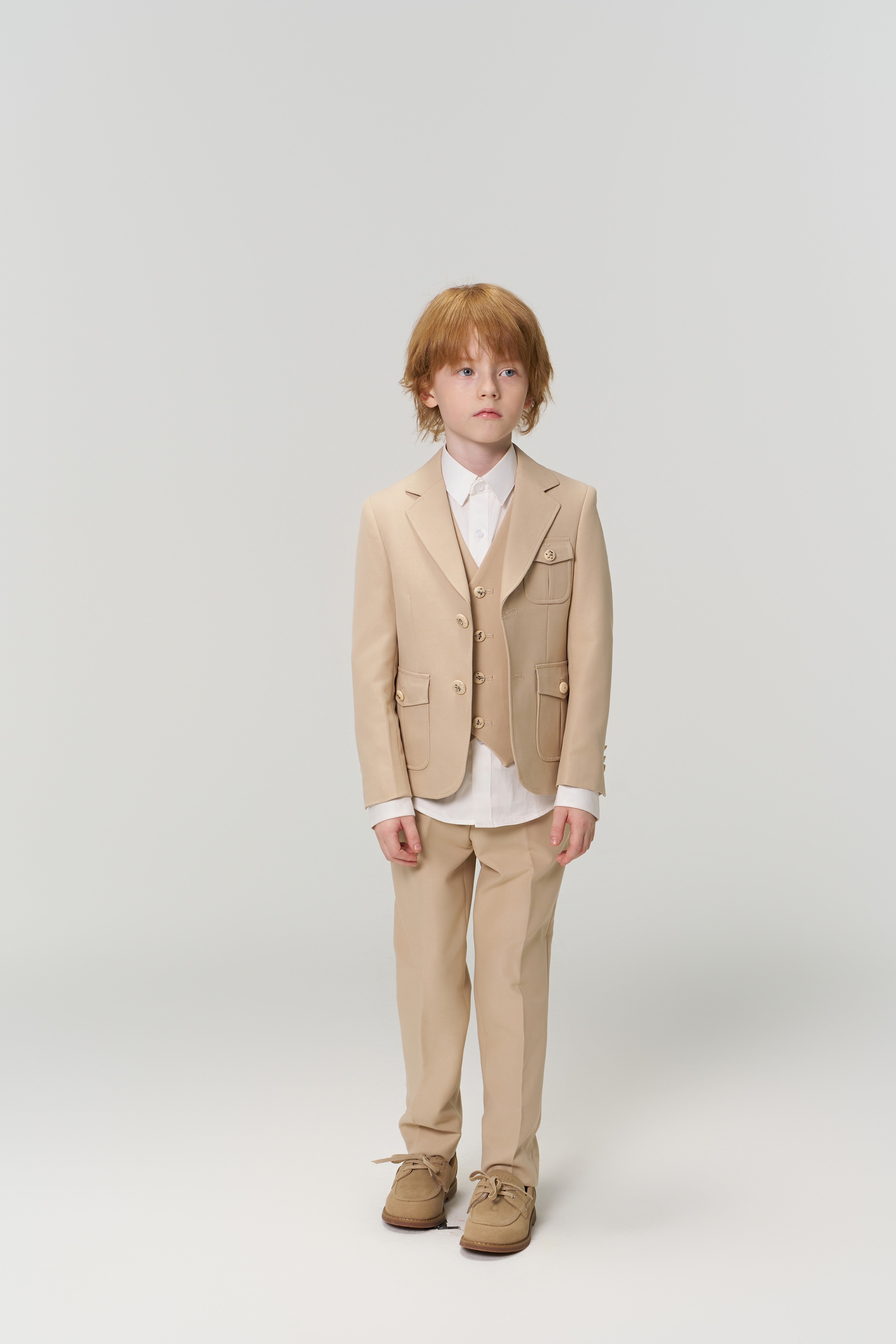 A young boy with red hair stands against a plain background, dressed for a special occasion in the KIKI and JOJO Savoy Dune Suit Vest, paired with a white shirt and brown shoes.