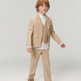 A young boy with light brown hair wears the KIKI and JOJO Savoy Dune Suit Vest over a white shirt with brown shoes, standing against a plain light background—ideal for any special occasion.