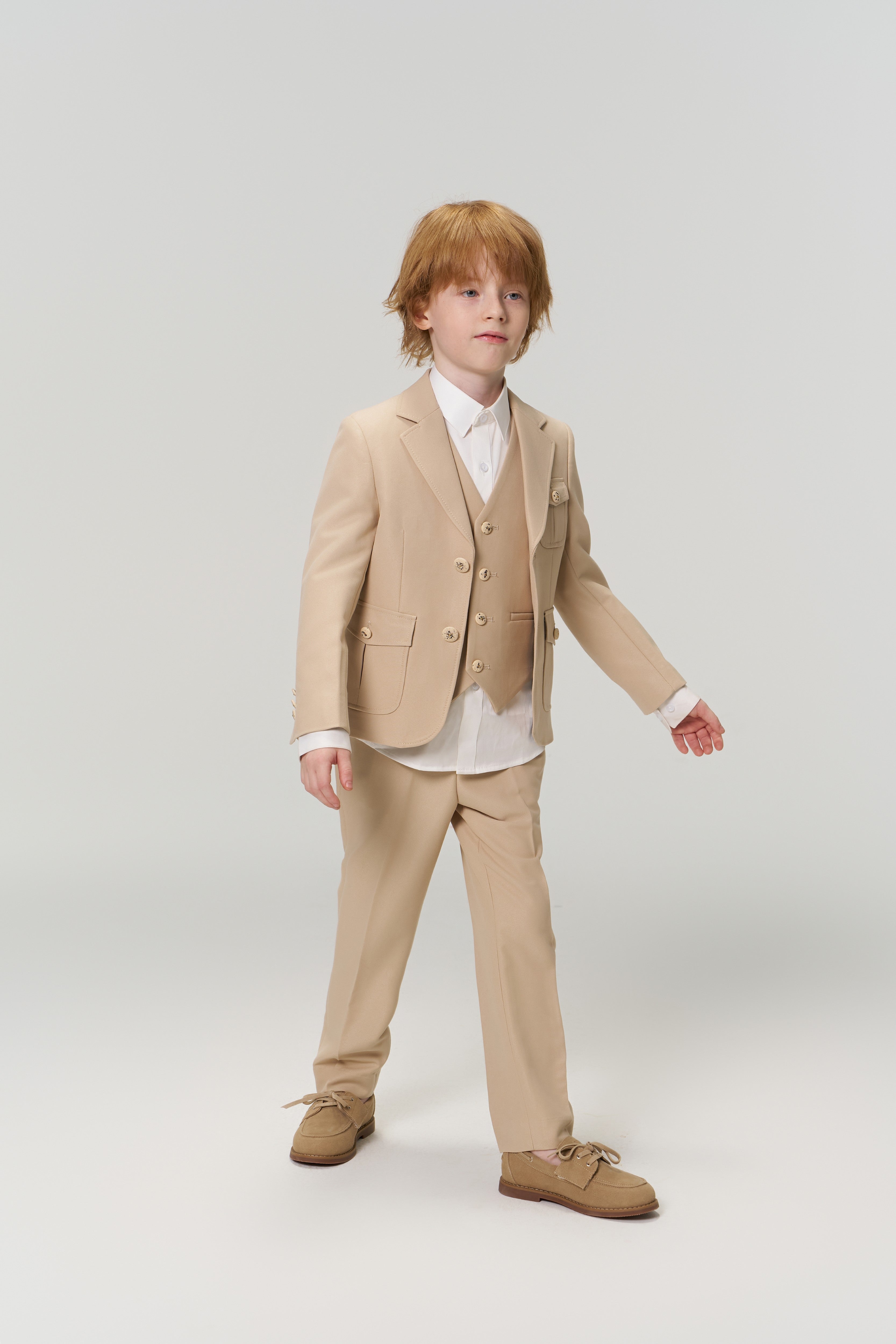 A young boy with light brown hair wears the KIKI and JOJO Savoy Dune Suit Vest over a white shirt with brown shoes, standing against a plain light background—ideal for any special occasion.