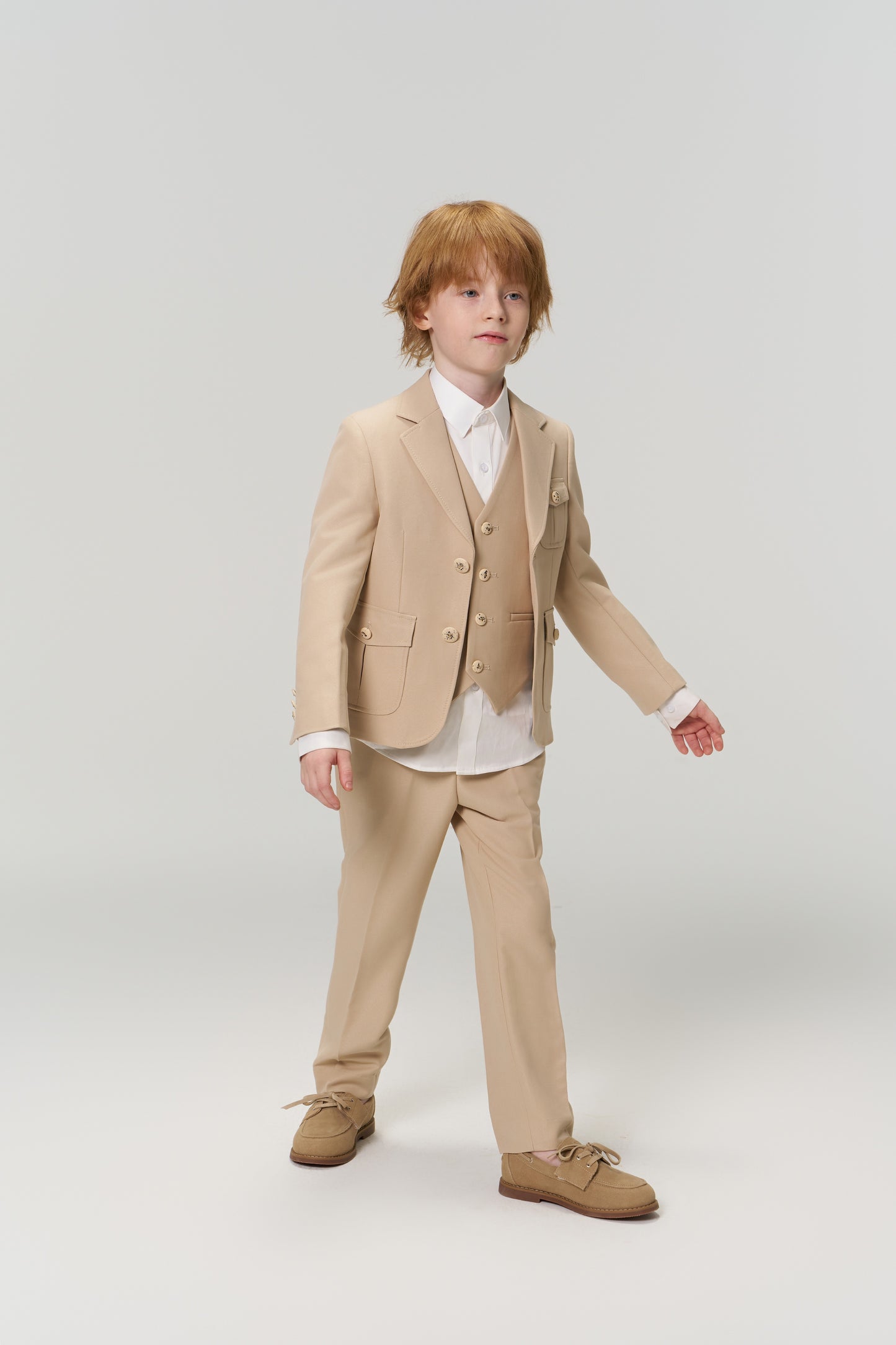 A young boy with light brown hair wears the KIKI and JOJO Savoy Dune Suit Vest over a white shirt with brown shoes, standing against a plain light background—ideal for any special occasion.