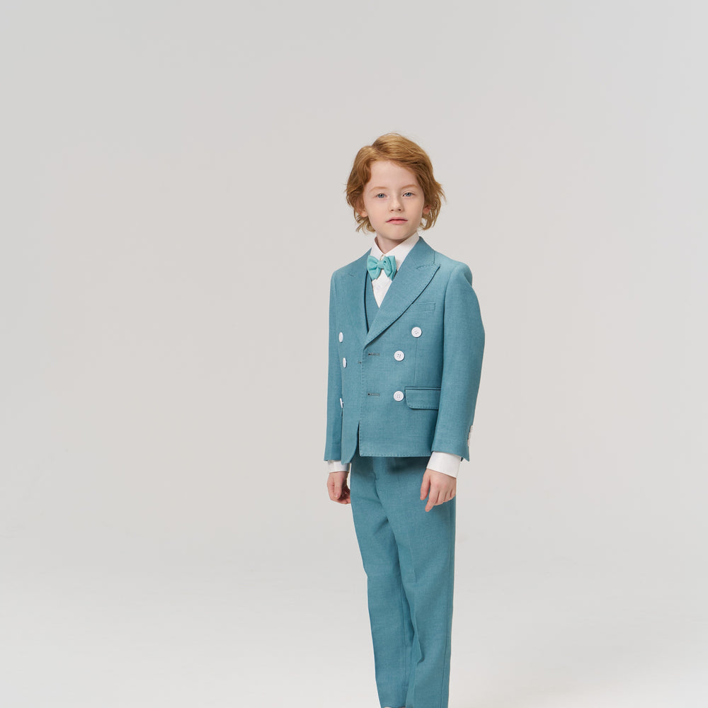 A young child with medium-length hair stands facing forward, wearing a light blue suit with white buttons, a tie, and white sneakers against a plain light background.