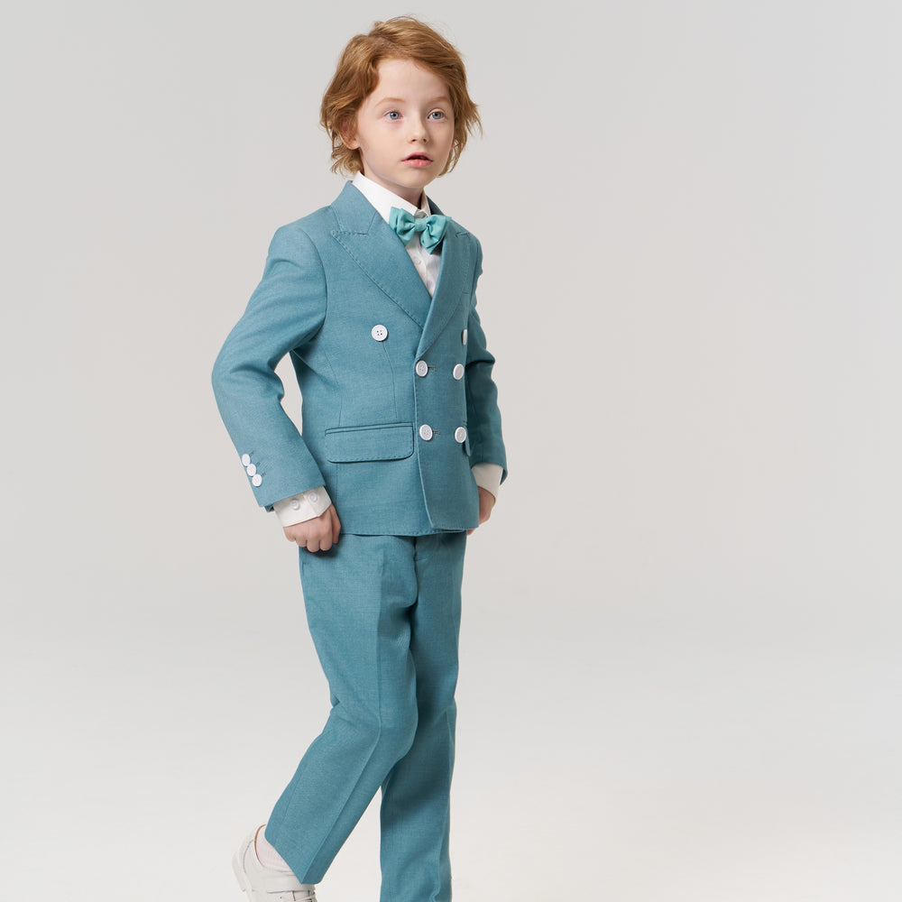 Young child with light brown hair stands in a light blue suit, white shirt, and bow tie against a plain white background.