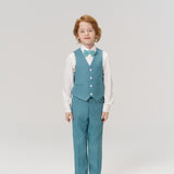 A young boy stands facing forward, wearing the KIKI and JOJO Celeste Whimsy Suit Vest with matching pants in peacock blue, a white shirt, bow tie, and white shoes against a plain light background.
