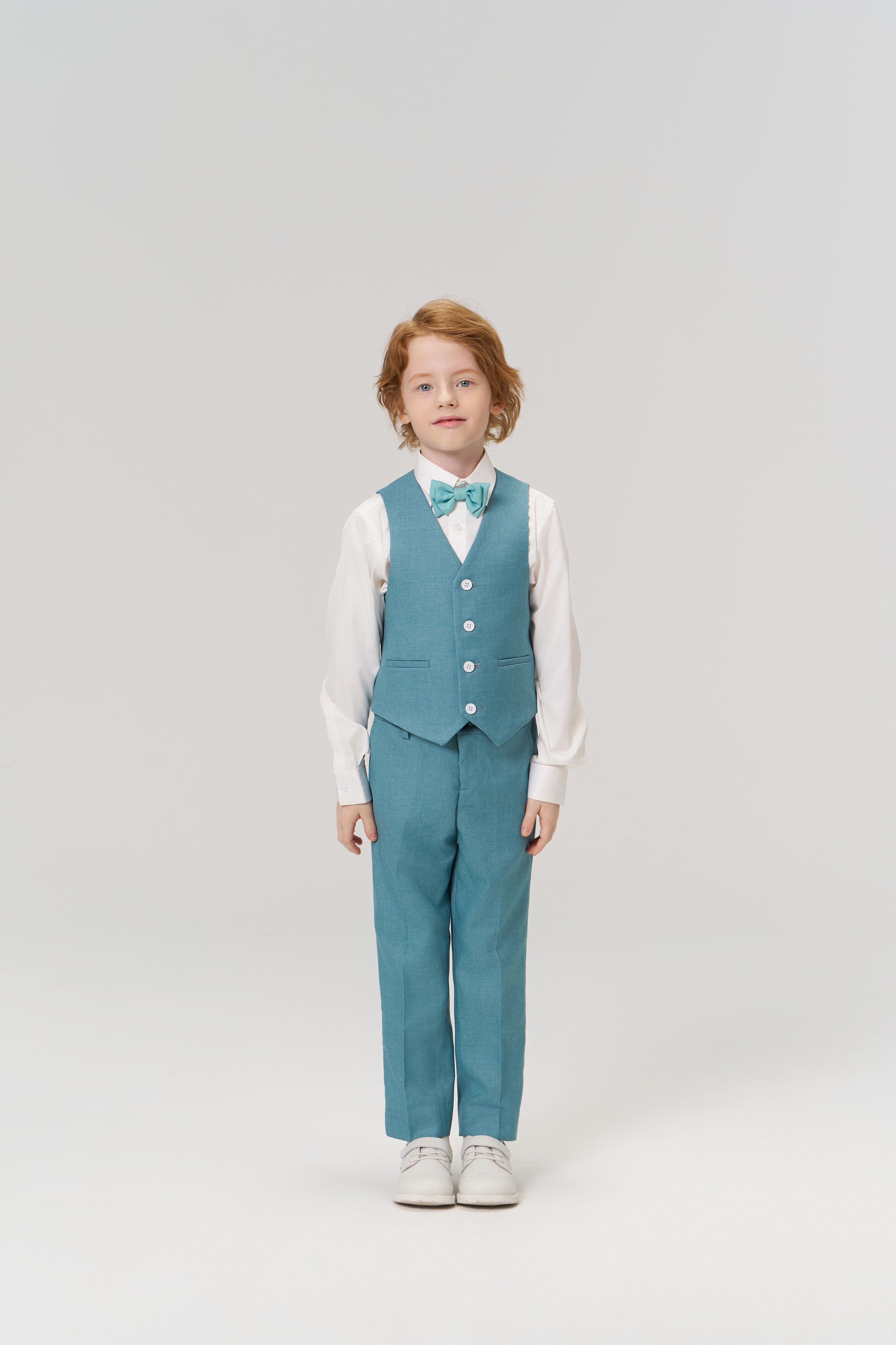 A young boy stands facing forward, wearing the KIKI and JOJO Celeste Whimsy Suit Vest with matching pants in peacock blue, a white shirt, bow tie, and white shoes against a plain light background.