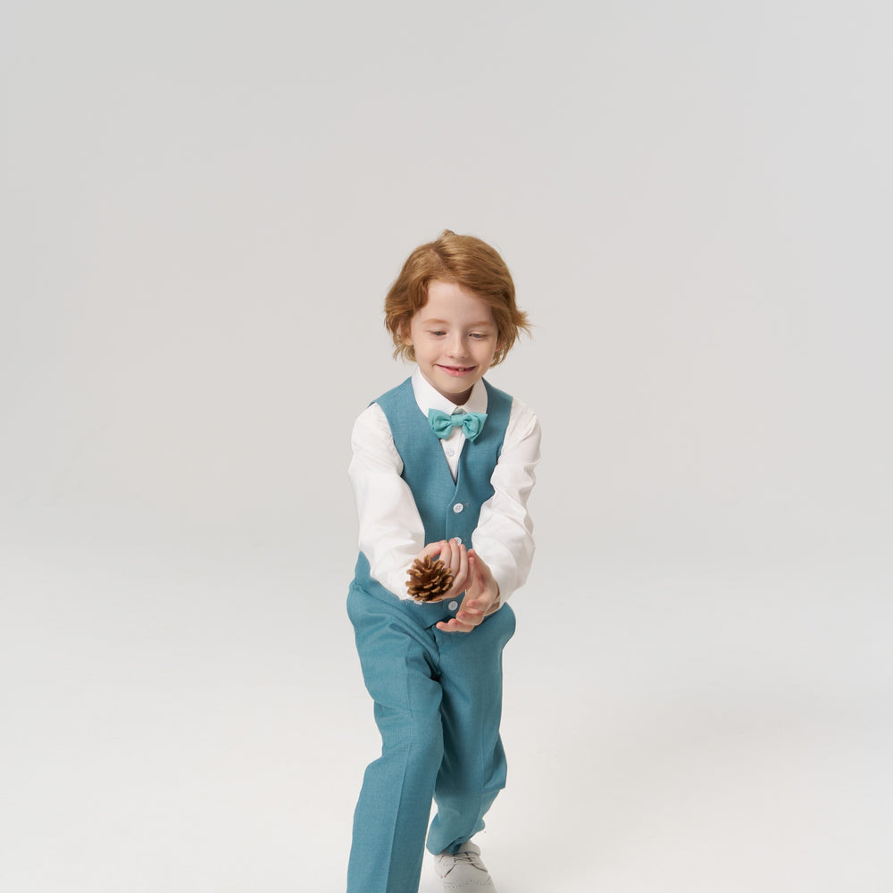 A young boy in a teal vest and bow tie tosses a pine cone, standing on a plain white background.