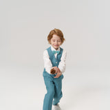 A young boy in a teal vest and bow tie tosses a pine cone, standing on a plain white background.