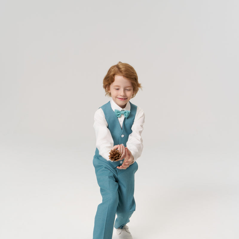 A young boy in a teal vest and bow tie tosses a pine cone, standing on a plain white background.